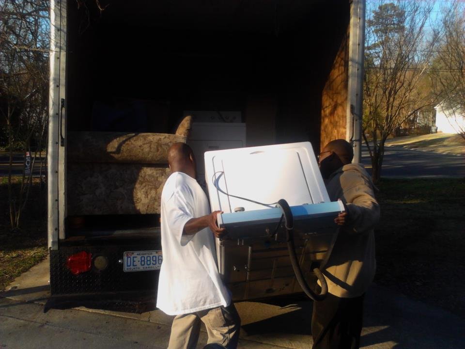 Two movers carrying a washing machine into a moving truck, with a couch visible in the truck's interior, representing professional moving services by Aim 2 Please Moving in North Carolina.