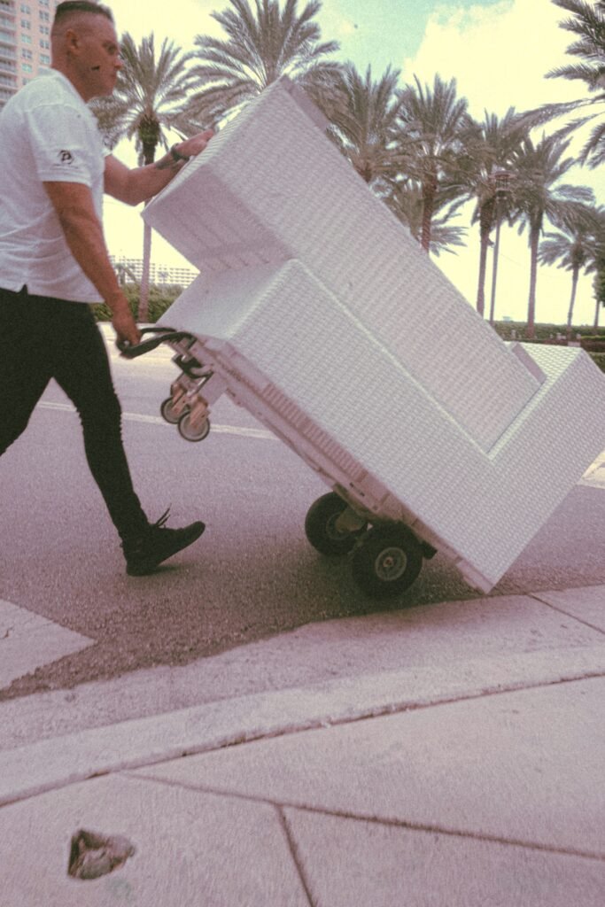 Mover transporting a white furniture piece on a dolly through a palm-lined street, emphasizing professional moving services by Aim 2 Please Moving.